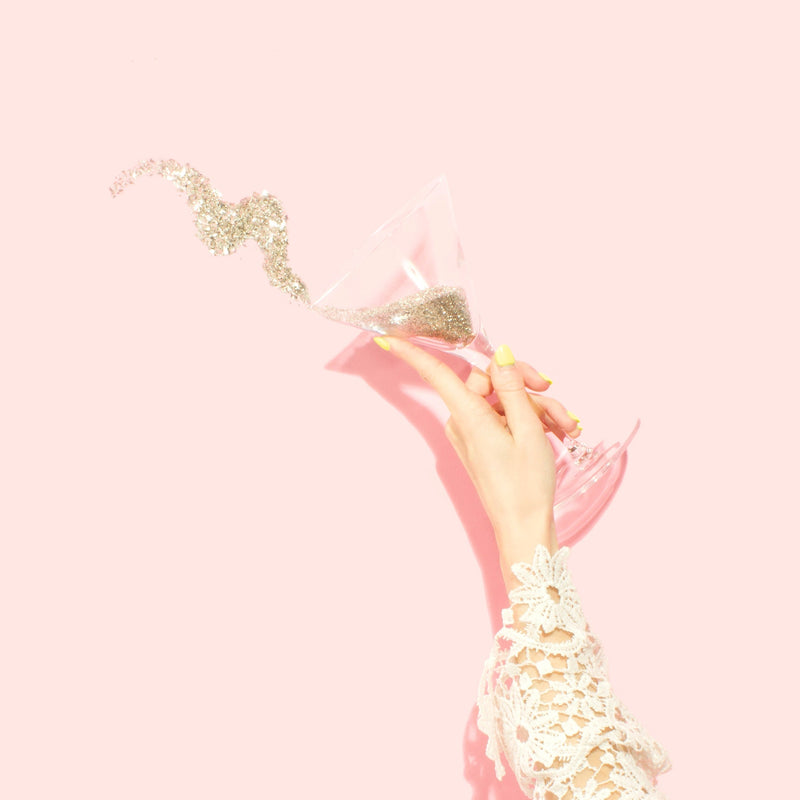 A hand holding a champagne flute with sparkling glitter spilling out against a pink background.