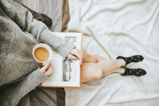 Person sitting on a bed wearing a gray sweater and black socks, holding a cup of coffee and an open book, illustrating enjoying quiet moments and mindfulness.