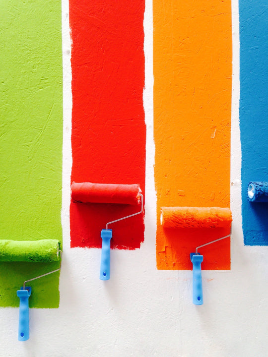 Three paint rollers applying vibrant colors: green, orange, and red on a wall.