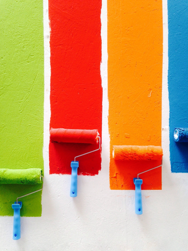 Three paint rollers applying vibrant colors: green, orange, and red on a wall.
