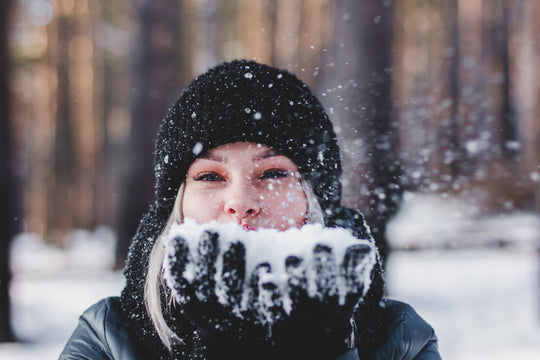 Woman in a black knit hat blowing snow from her gloved hands in a snowy forest