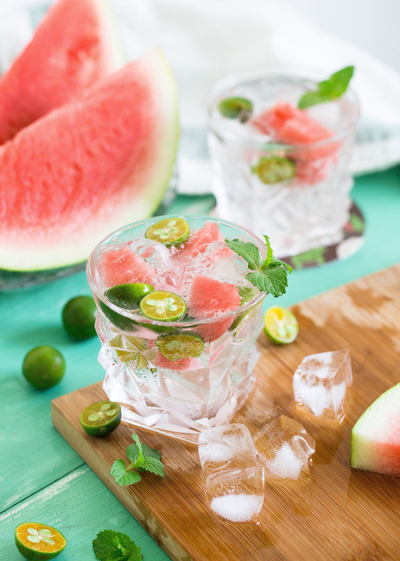 Glass of watermelon-infused water with lime slices, mint, and ice on a wooden board, watermelon slices in the background
