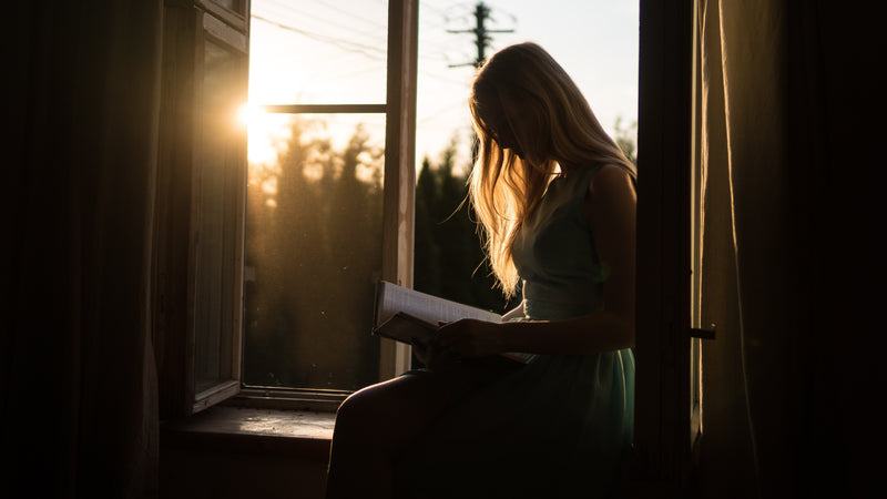Silhouette of a woman reading a book while sitting on a windowsill with sunlight streaming through the window at sunset.