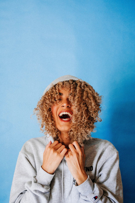 Person with curly blonde hair wearing a gray hoodie laughing against a blue background
