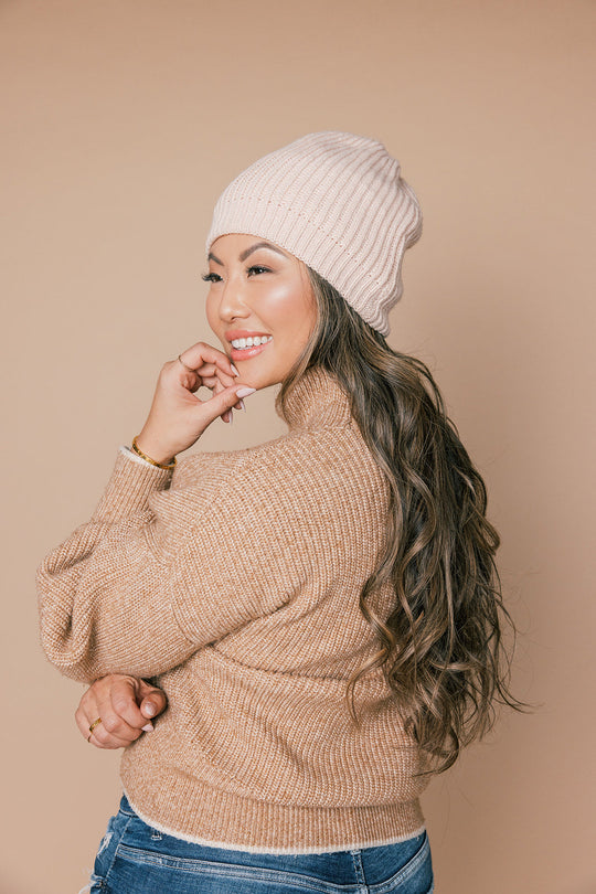 Smiling woman touching her chin wearing a pink knit beanie and beige sweater, showing long wavy hair against a beige background