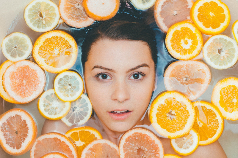 A woman with dark hair submerged in water surrounded by floating slices of citrus fruits including oranges, lemons, and grapefruits, symbolizing natural skincare ingredients.