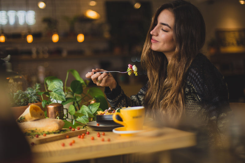 Woman enjoying a meal at a wooden table with plants and a yellow coffee cup in a cozy cafe setting