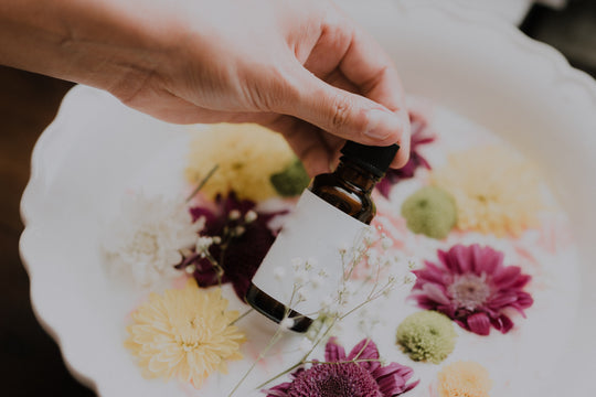 Hand holding a small amber essential oil bottle with a blank white label over a white bowl filled with colorful flowers including yellow, purple, and white blossoms.