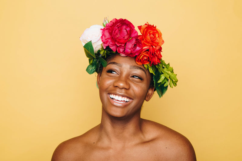 A woman smiling brightly, wearing a colorful flower crown against a yellow background.