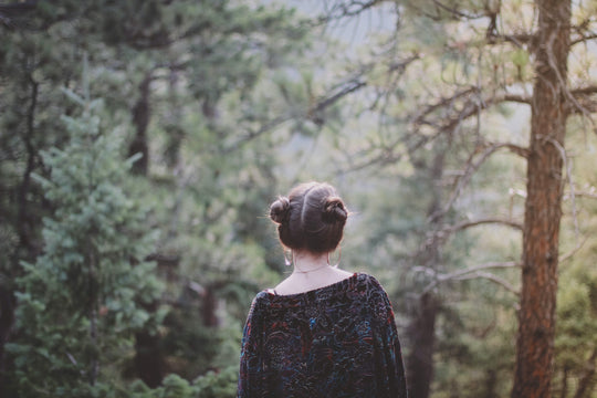 Woman with hair in two buns seen from behind standing among pine trees in a sunlit forest.