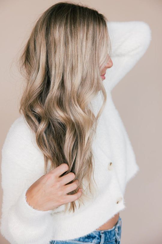 Woman side profile touching long wavy blonde hair against a neutral background, wearing a white sweater