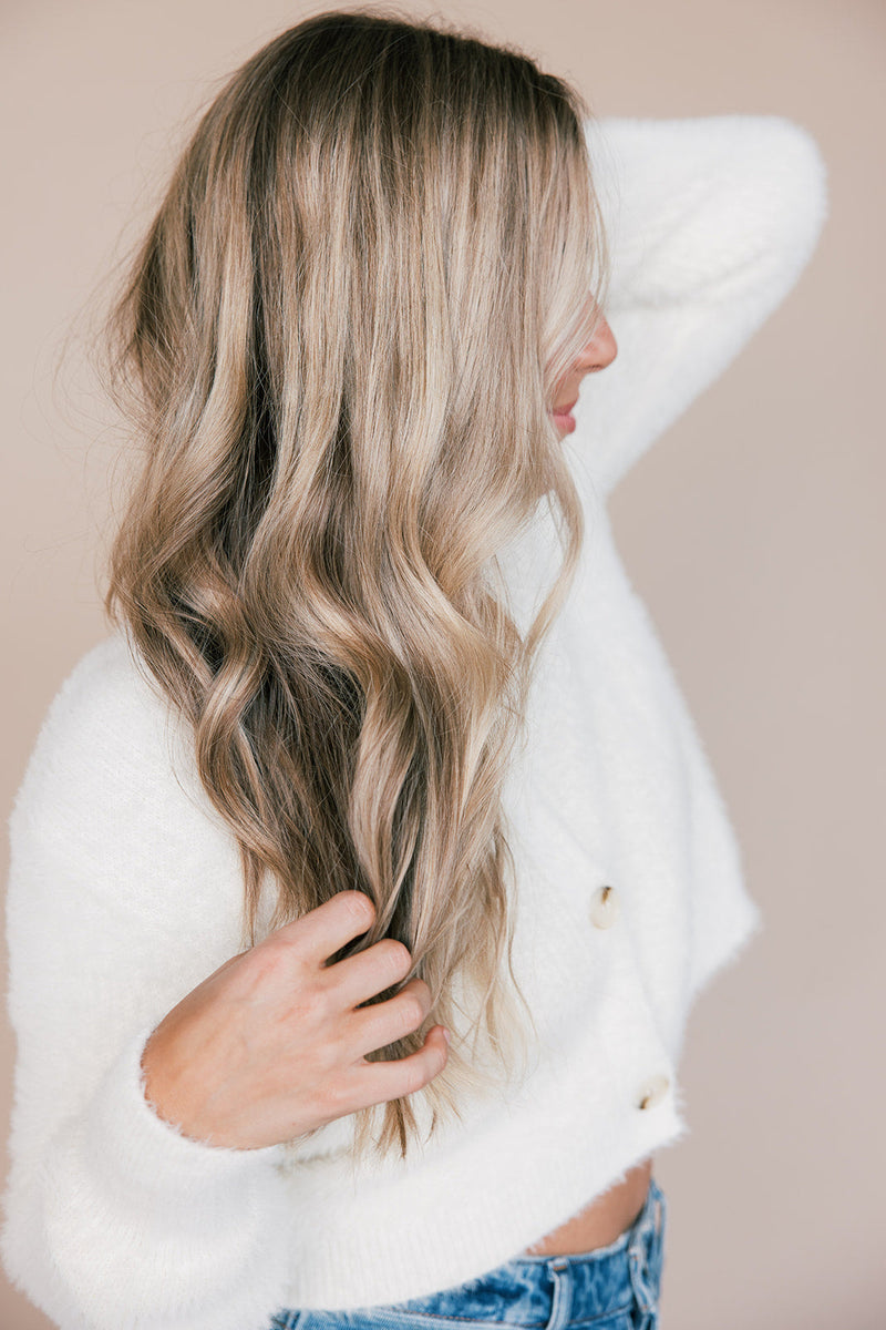 Woman side profile touching long wavy blonde hair against a neutral background, wearing a white sweater