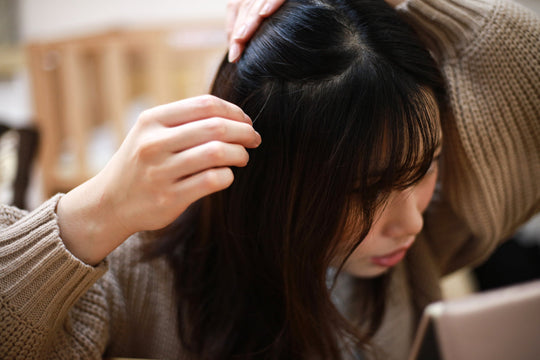 Woman parting her dark hair to inspect her scalp while leaning over a mirror in a cozy indoor setting