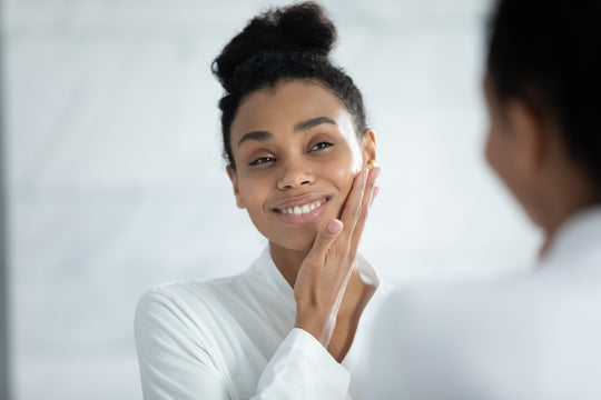Woman smiling and gently touching her cheek in front of a bathroom mirror, wearing a white robe