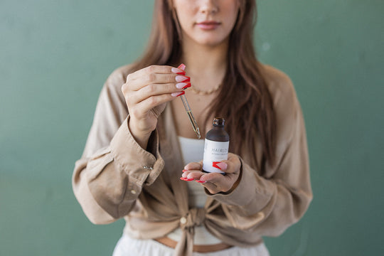 Front view of HAIRLOVE Growth Complex dropper bottle held by woman, dropper lifted above bottle against soft green background