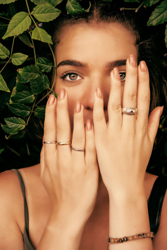 Woman holding her hands over her face to show manicured nails and rings against a leafy background