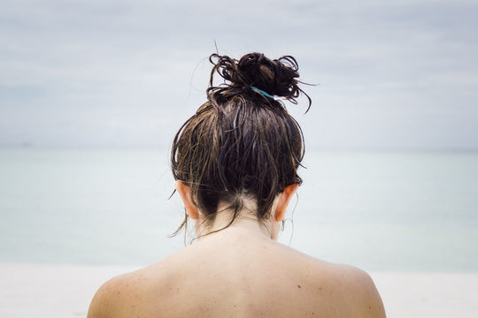 Woman with wet hair in a messy bun facing the ocean on a beach, bare shoulders visible