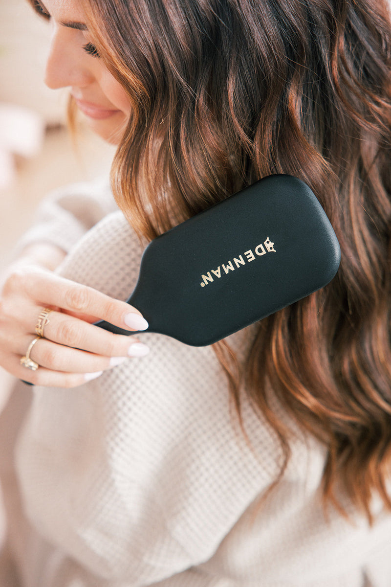 Woman brushing long brown hair with a black Denman hair brush showing the word 'DENMAN' in gold letters