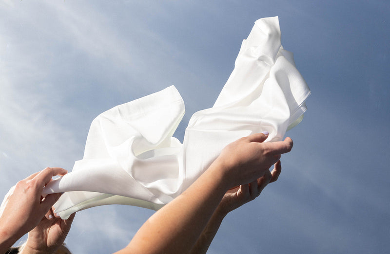 Hands holding a white silk or satin fabric against a blue sky background.