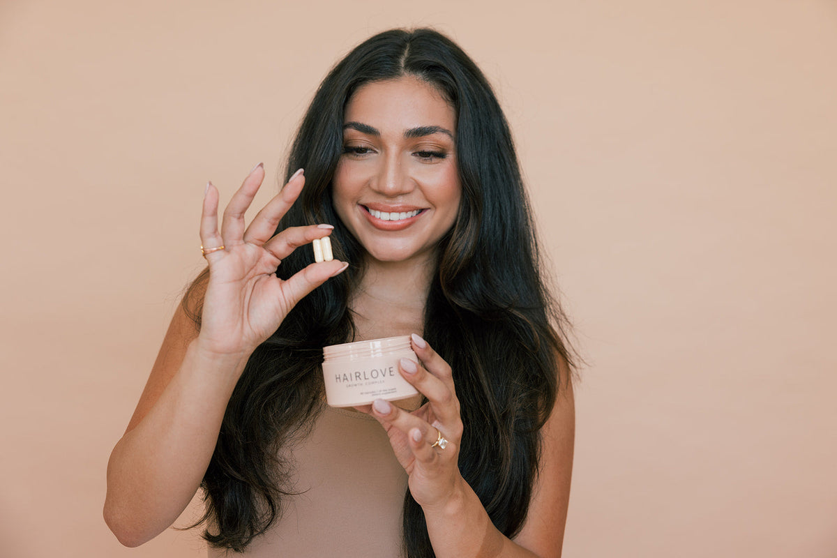 A smiling woman with long dark hair holding two HAIRLOVE capsules in one hand and a HAIRLOVE Growth Complex container in the other.