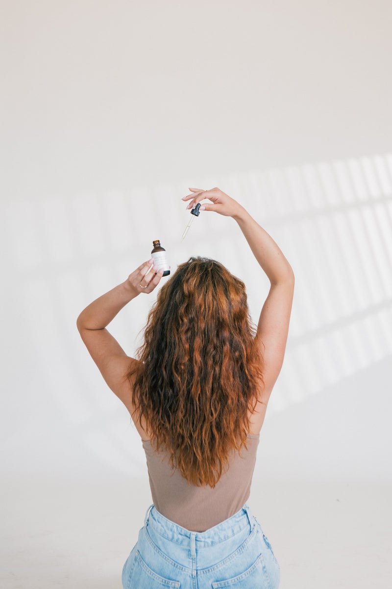 Woman with long wavy hair seen from behind applying scalp treatment with a dropper from a brown bottle against a plain light background.