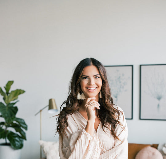 Smiling woman with long wavy brown hair standing in a bright living room, wearing a cream blouse and tassel earrings