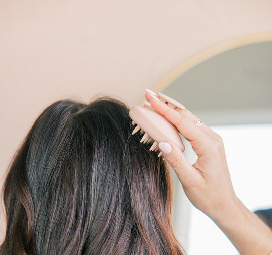 Person using a pink handheld scalp massager on dark brown hair in front of a mirror