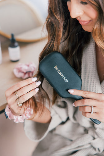 A woman brushing her hair with a Denman brush while wearing a robe, with hair products in the background.