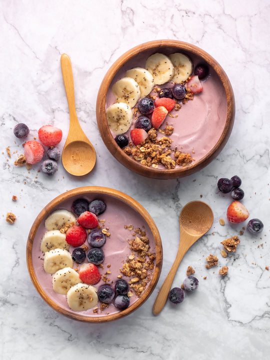 Two purple smoothie bowls topped with banana slices, strawberries, blueberries, chia seeds and granola on marble with wooden spoons