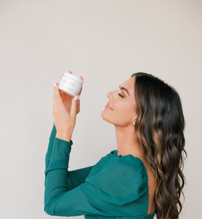 Woman with long dark hair in a green top holding and looking at a HAIRLOVE Growth Complex jar with visible text 'HAIRLOVE' on the label.