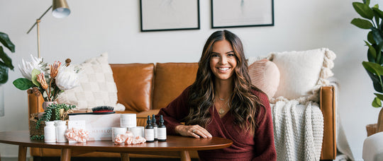 Smiling woman with long brown hair seated at a table displaying hair-care products in a cozy living room