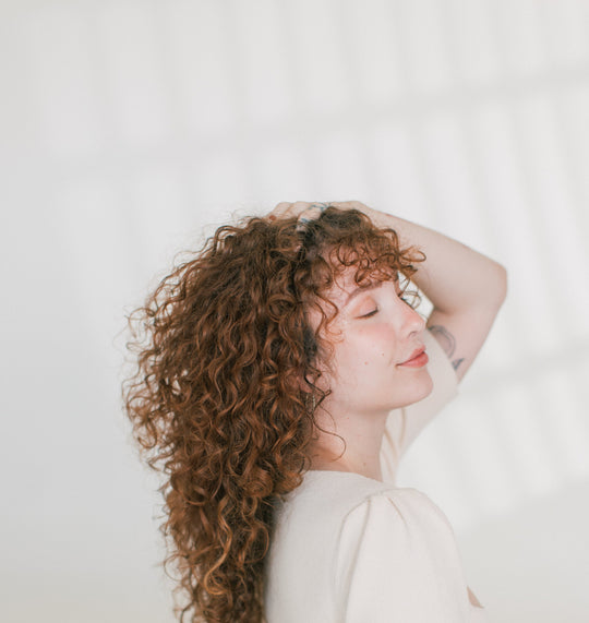 Woman with curly brown hair touching her hair and smiling with eyes closed against a soft white background