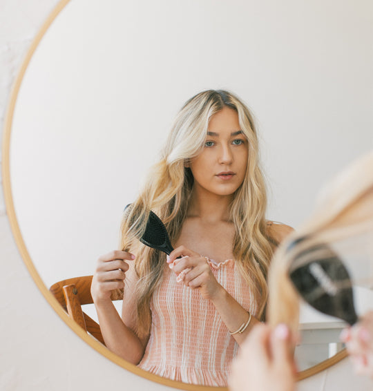 Young blonde woman brushing long wavy hair while seated at a wooden chair, reflected in a round mirror against a neutral wall