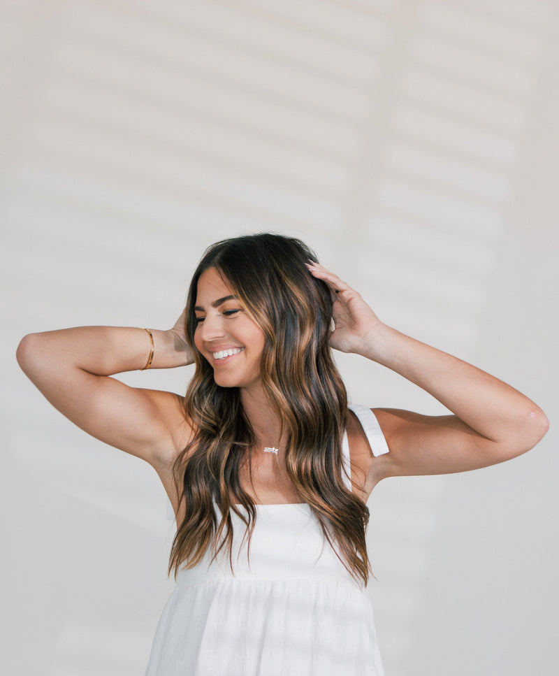 Smiling woman with long wavy brown hair wearing a white sleeveless top, posing with hands behind her head against a light background