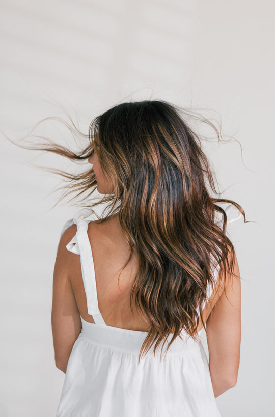 Woman turning away with long wavy brown hair and caramel highlights blowing in the wind against a light backdrop