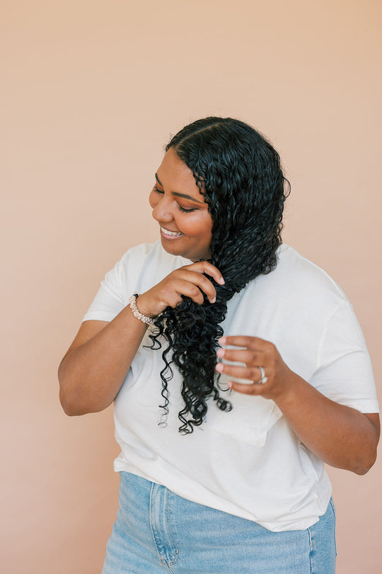Smiling person with long, curly black hair wearing a white t-shirt and blue jeans, touching their hair against a beige background