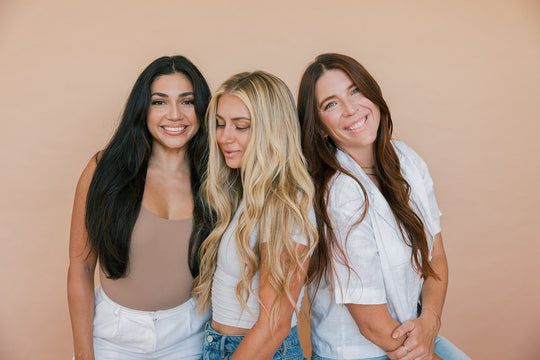 Three women with long black, blonde, and auburn hair smiling together against a warm beige backdrop
