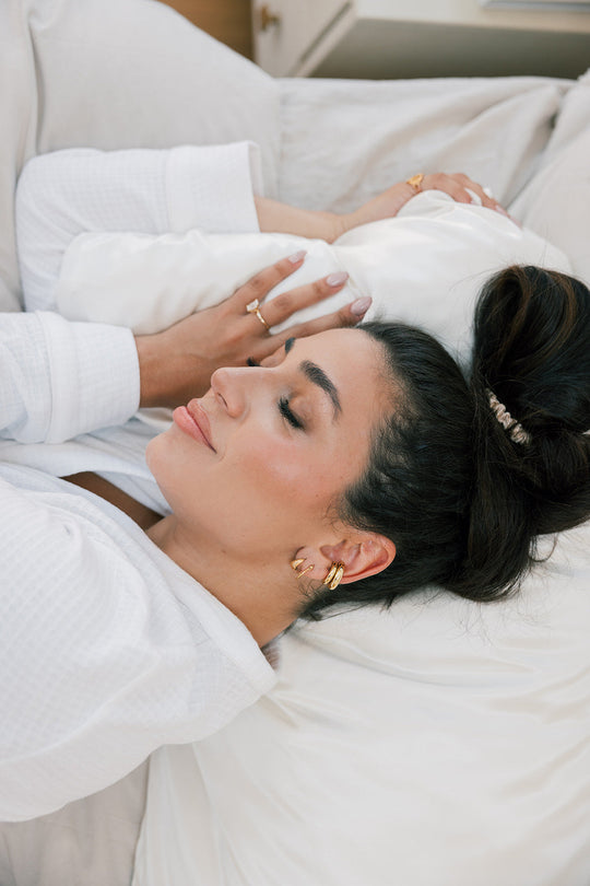 Woman lying on a bed with eyes closed, hair in a loose bun, wearing gold earrings and a white robe, resting on a pillow