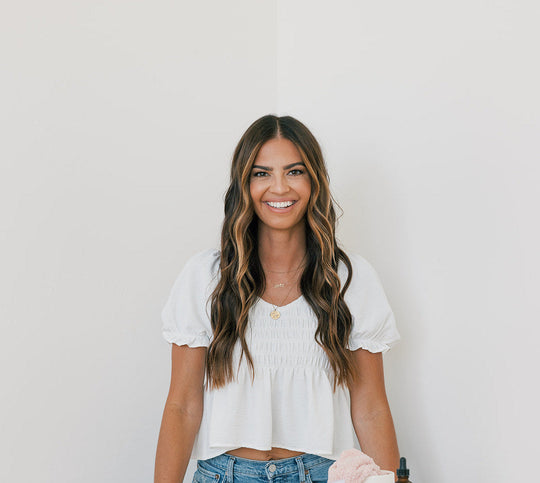 Smiling woman with long wavy brown hair wearing a white puff-sleeve top and blue jeans, standing against a plain light background, holding hair care products.