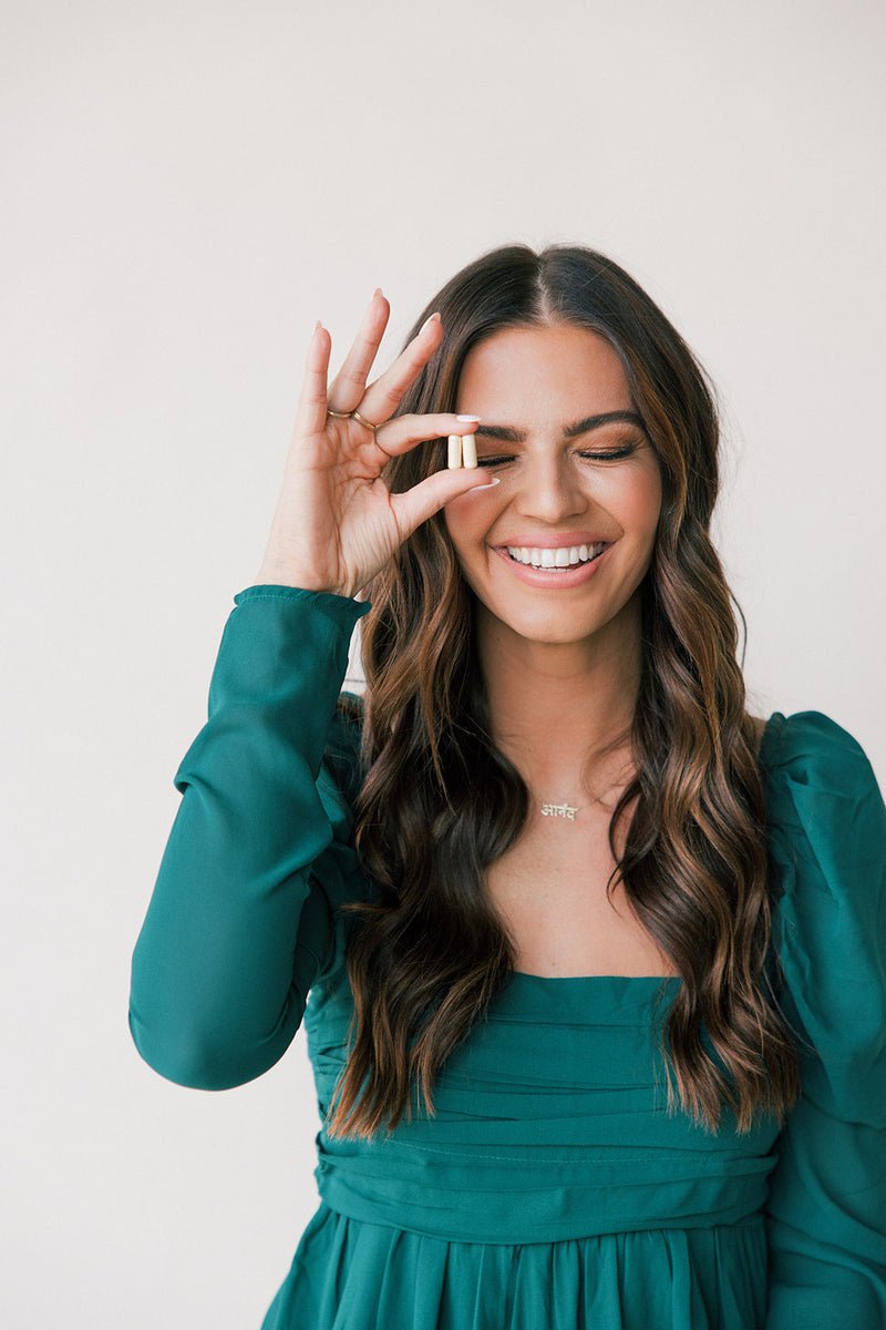 Smiling woman in a teal dress holding two HAIRLOVE Growth Complex capsules near her eye against a plain white background