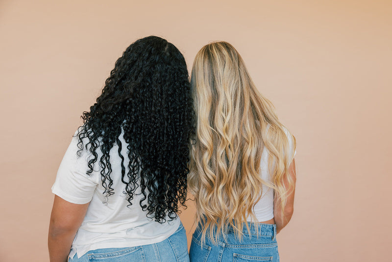 Two women with their backs to the camera showing long curly black hair and long wavy blonde hair against a peach background