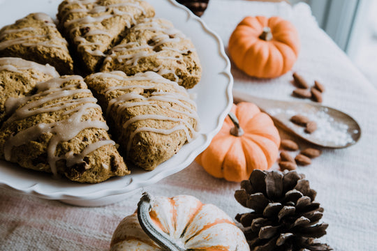Iced scones drizzled with icing on a white pedestal plate, surrounded by mini orange and white pumpkins, a pinecone, and almonds on a linen table