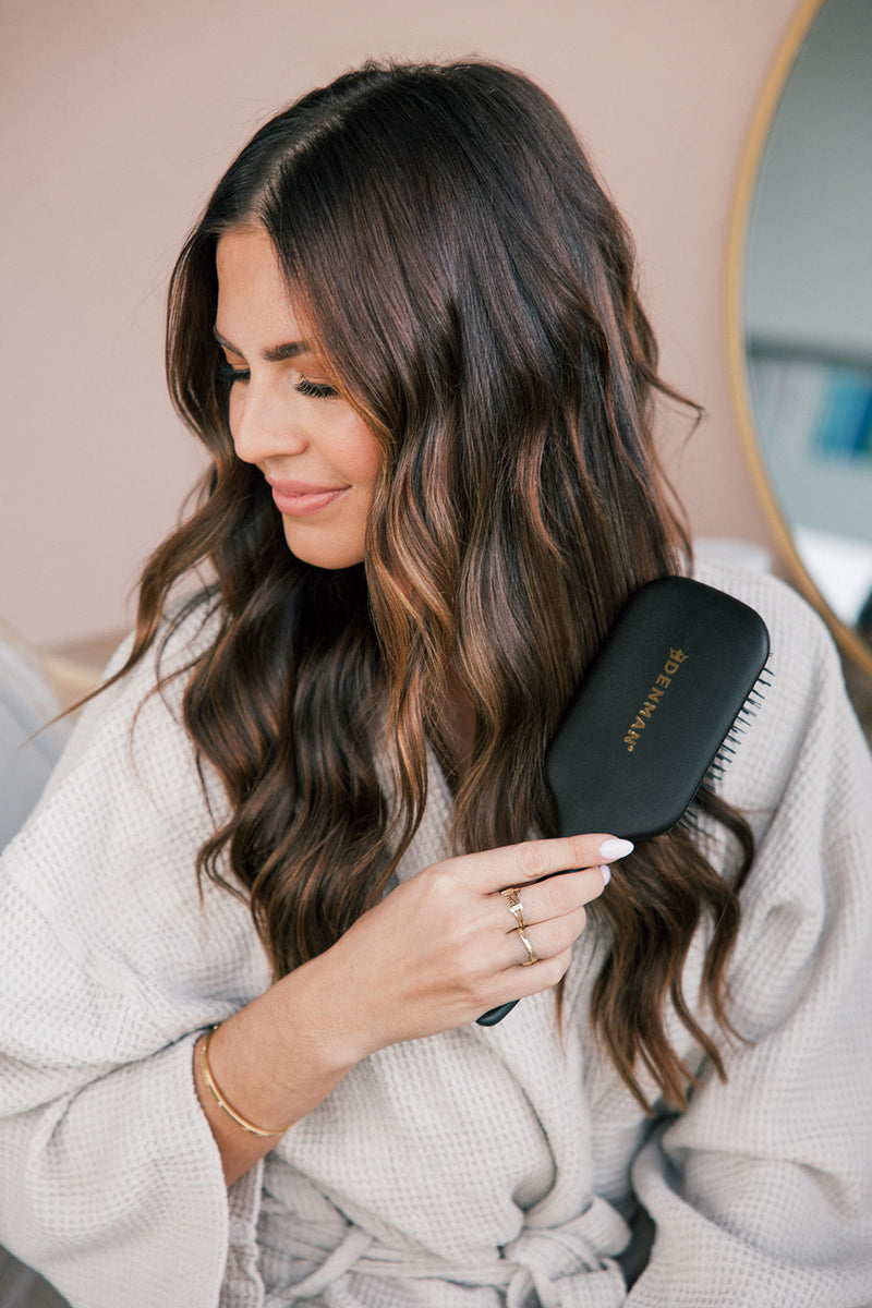 Woman brushing her long wavy brown hair with a black Denman hairbrush, wearing a light-colored robe, smiling gently.
