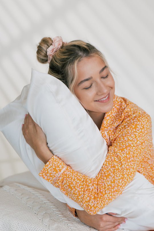 Young woman hugging a white pillow and smiling peacefully while sitting on a bed in orange floral pajamas