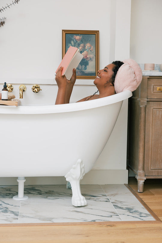 Woman with a pink towel wrapped around her hair, smiling and reading a book while relaxing in a white clawfoot bathtub in a cozy bathroom setting.