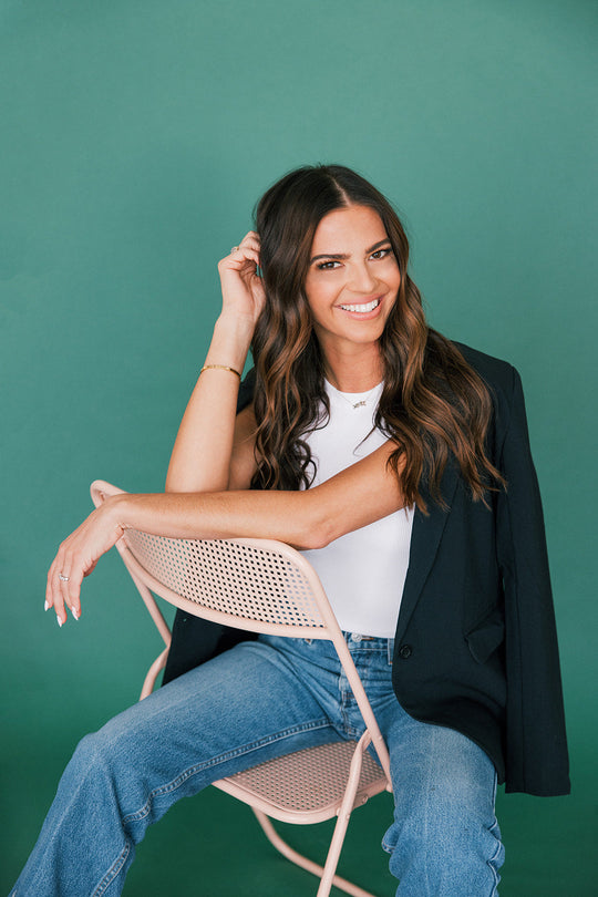 Woman with long wavy brown hair smiling, seated on a pink chair against a green backdrop wearing a white top and jeans