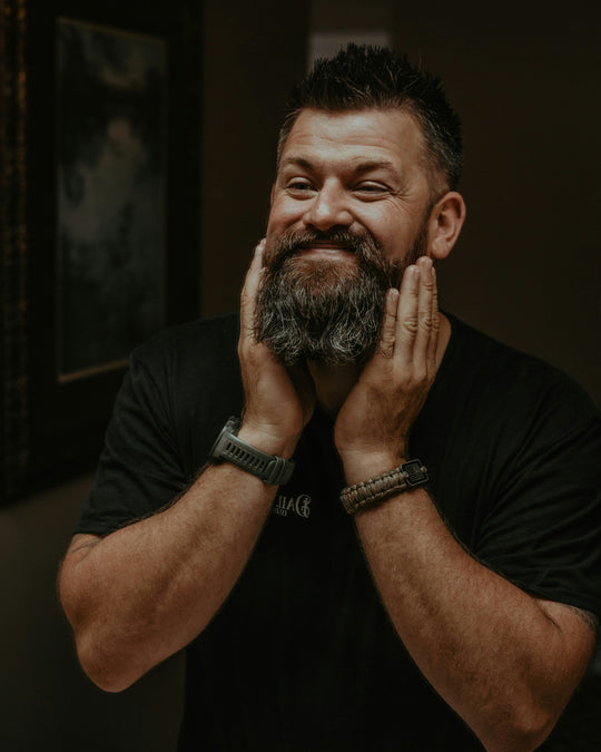 Smiling man with a full beard touching his beard with both hands, wearing a black shirt and wristbands, in a dimly lit room.