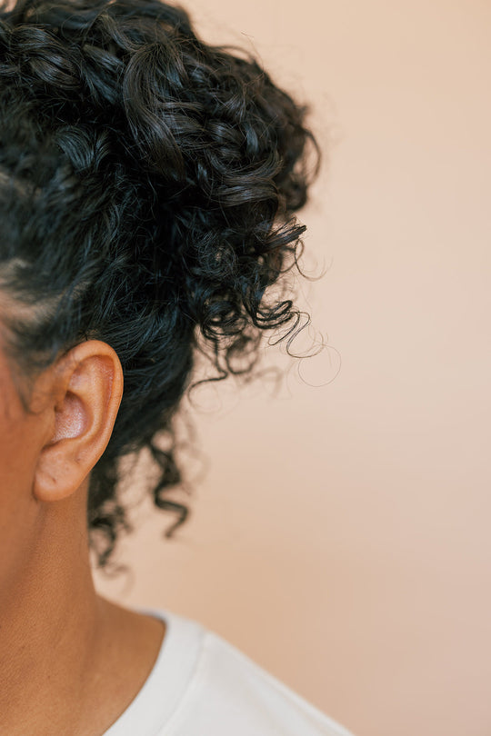 Close-up of a woman with curly black hair tied up, showing the side of her head and ear against a beige background.