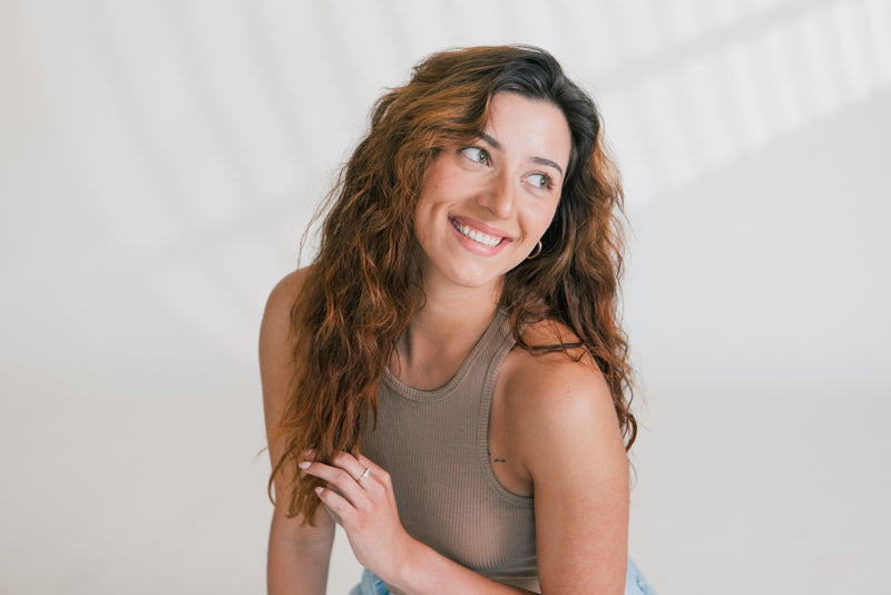 Smiling woman with long wavy hair and glowing skin wearing a sleeveless beige top against a light background