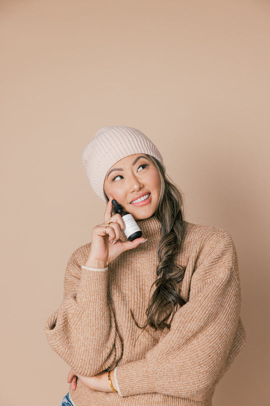 Woman in beige sweater and beanie holding HAIRLOVE Nourish and Repair Serum to her cheek, smiling against a beige background
